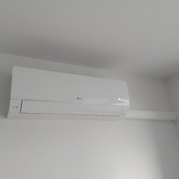 Breezo Piotr Wiśniewski - Biała klimatyzacja LG Dual Inverter zamontowana na białej ścianie, widoczny fragment rury odprowadzającej. Minimalistyczny design i czyste linie.