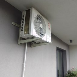 Breezo Piotr Wiśniewski - Zamontowany, biały klimatyzator Rotenso na szarej ścianie budynku, widok z dołu. Widoczne rury instalacyjne i fragment okna. Zewnętrzna jednostka klimatyzacji.