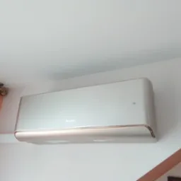 Klimatyzator Gree, model ścienny, zamontowany wewnątrz pomieszczenia. Widoczne detale montażu i wykończenia, biała obudowa z delikatnym, złotym akcentem.