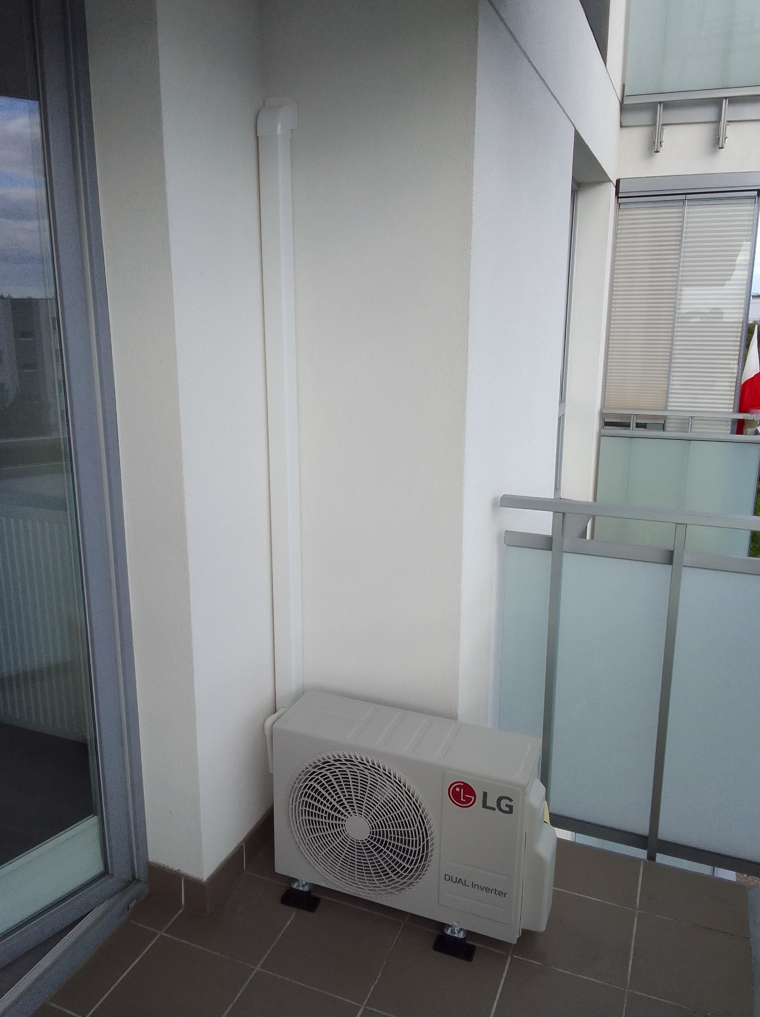 Biała jednostka zewnętrzna klimatyzacji LG Dual Inverter na balkonie, z widoczną rurą odprowadzającą. Tło stanowi balustrada i okno balkonowe.