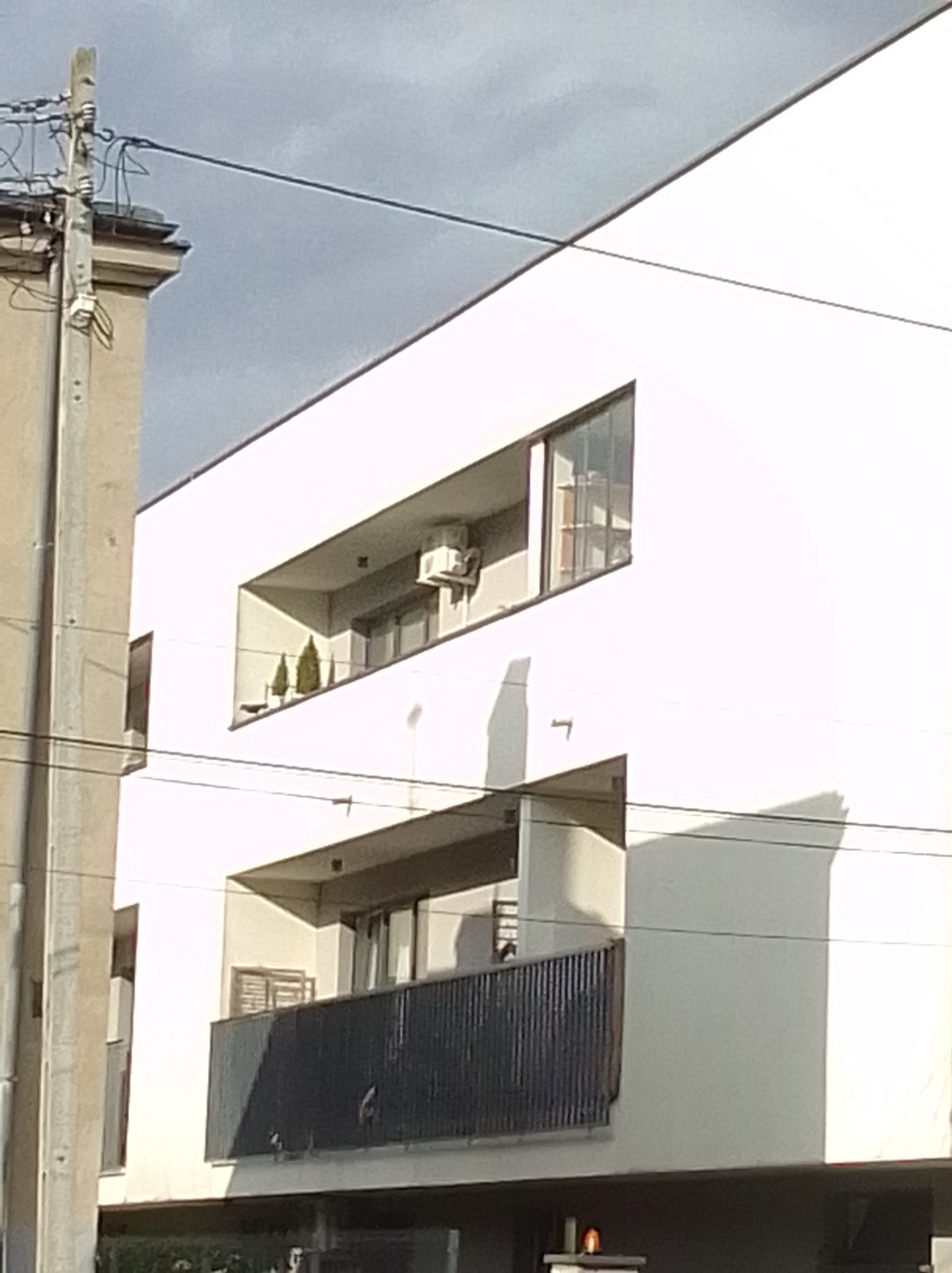 Biały budynek z balkonami; na jednym z nich widoczna jednostka zewnętrzna klimatyzacji, zieleń doniczkowa. Ujęcie z dołu, z widocznymi przewodami.