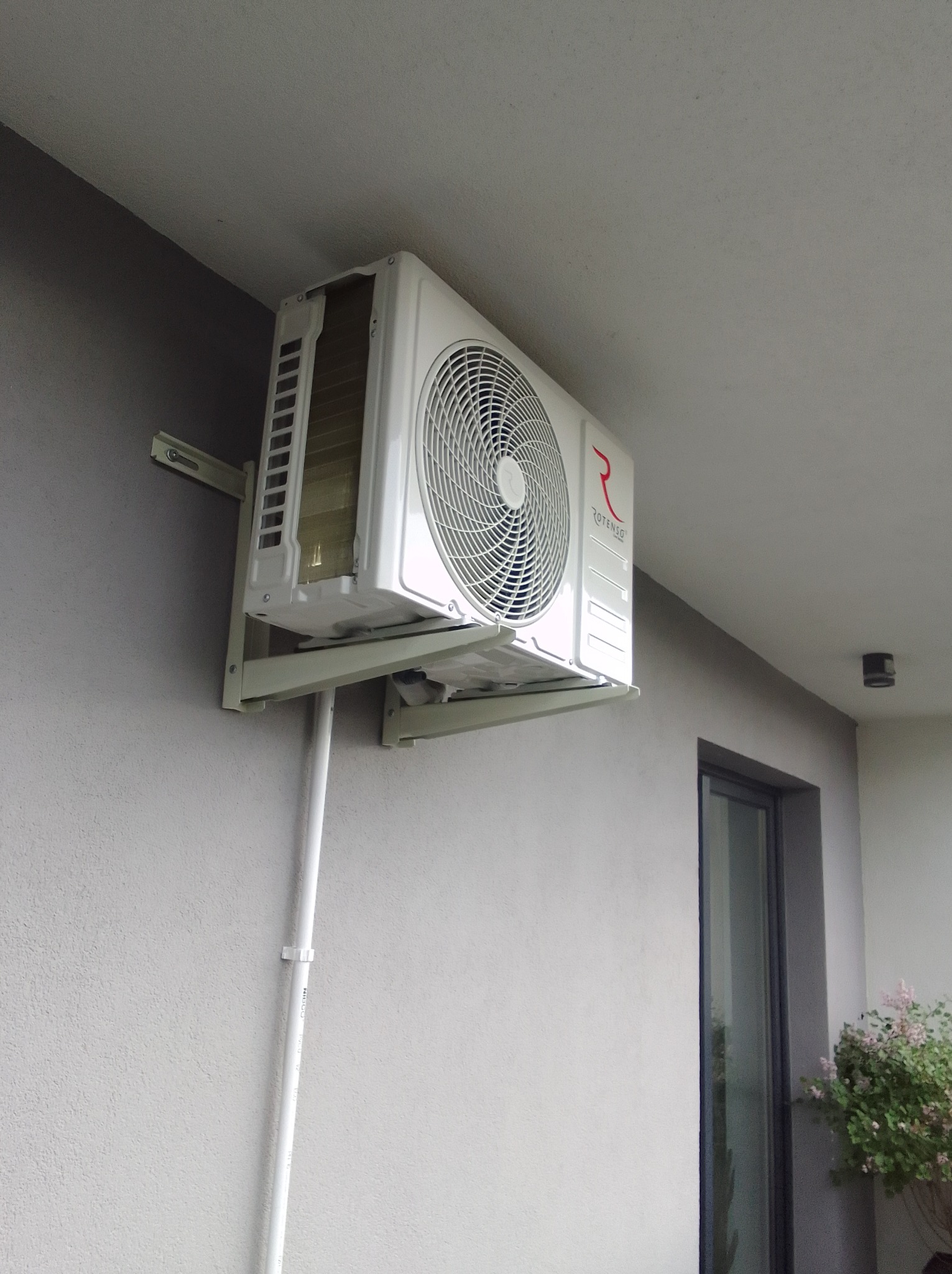 Zamontowany, biały klimatyzator Rotenso na szarej ścianie budynku, widok z dołu. Widoczne rury instalacyjne i fragment okna. Zewnętrzna jednostka klimatyzacji.