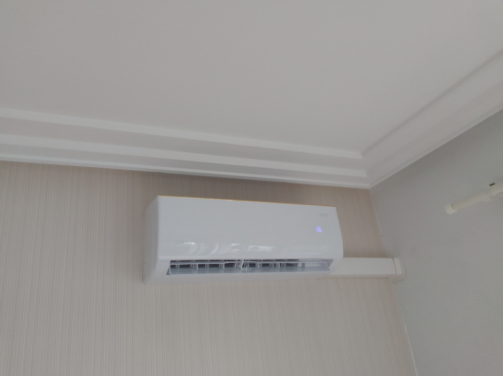 Biały klimatyzator ścienny marki Ronsu z wyświetlaczem temperatury 16 stopni, zamontowany w pomieszczeniu z jasną tapetą i ozdobnym sufitem.