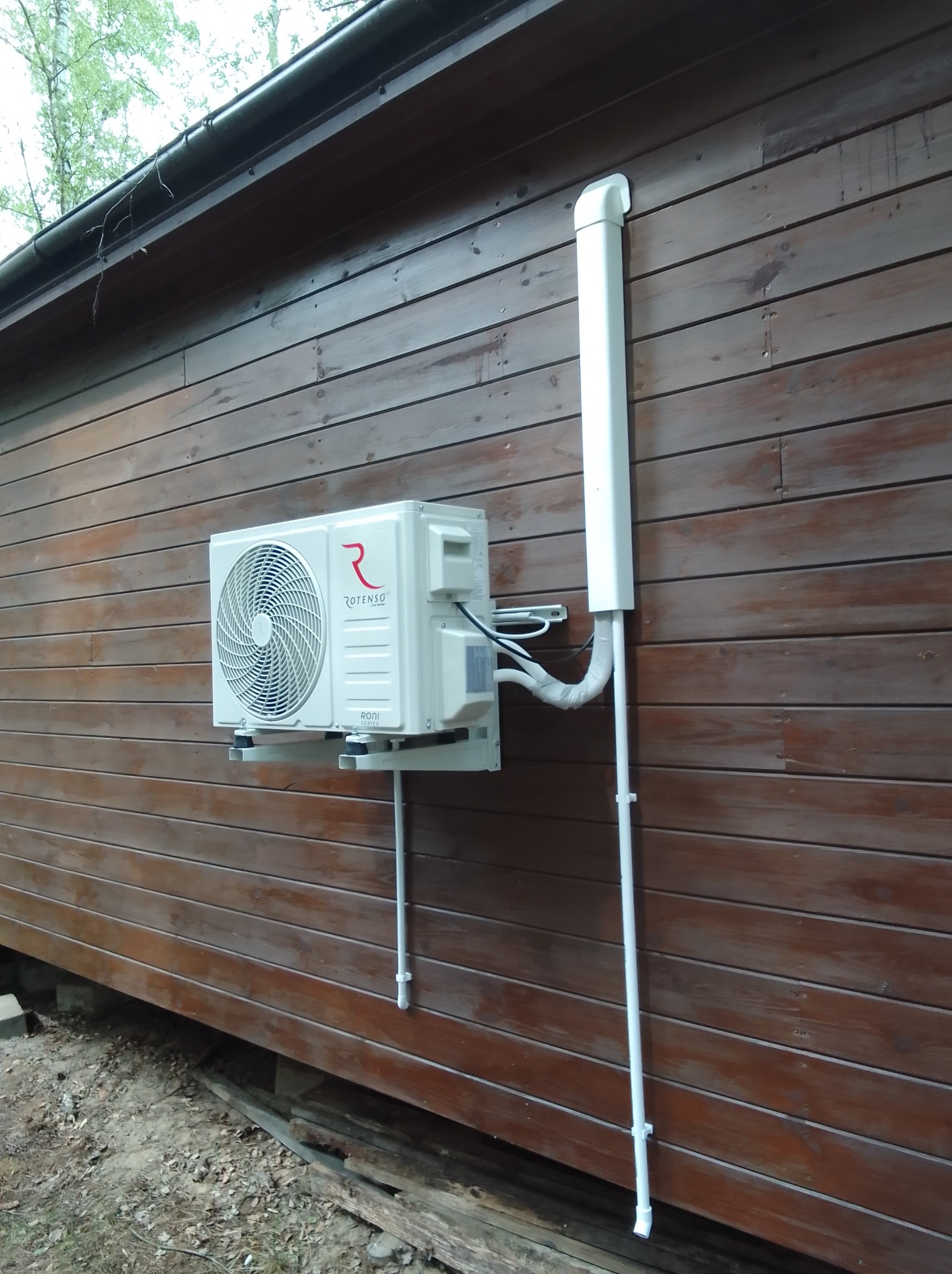 Biały klimatyzator Rotenso zamontowany na brązowej, drewnianej elewacji budynku. Widoczne rury odprowadzające i elementy montażowe.