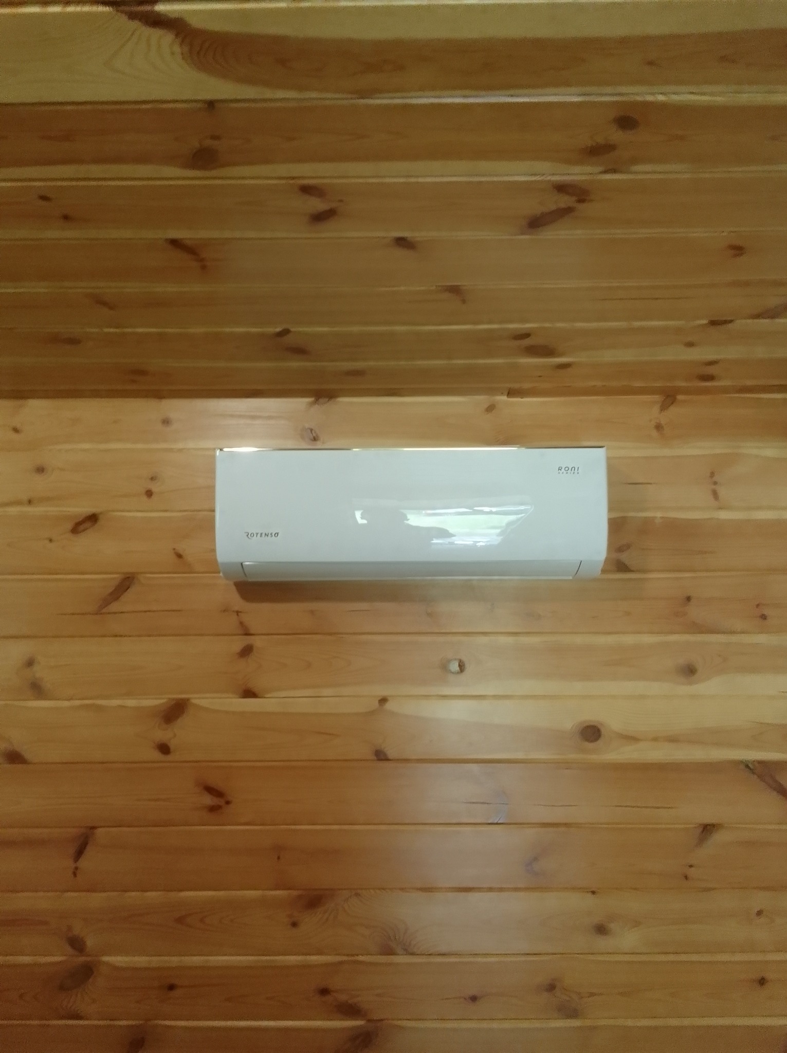 Biały klimatyzator Rotenso zamontowany na ścianie z drewnianych paneli, widok z dołu, ciepłe oświetlenie wnętrza.
