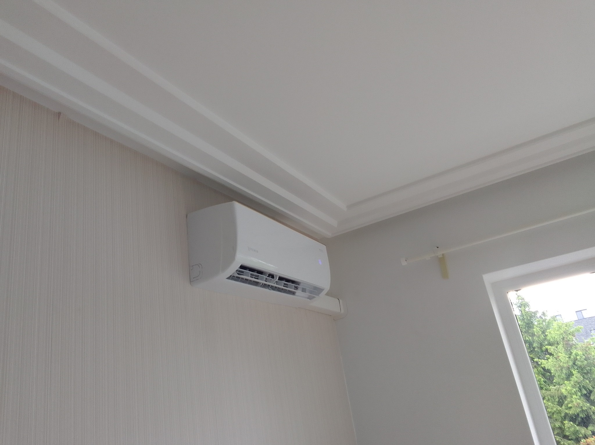 Biała klimatyzacja zamontowana na ścianie w Brańszczyku, widok z dołu. Urządzenie marki Rotenso, z widocznym wyświetlaczem. W tle okno z zielenią.