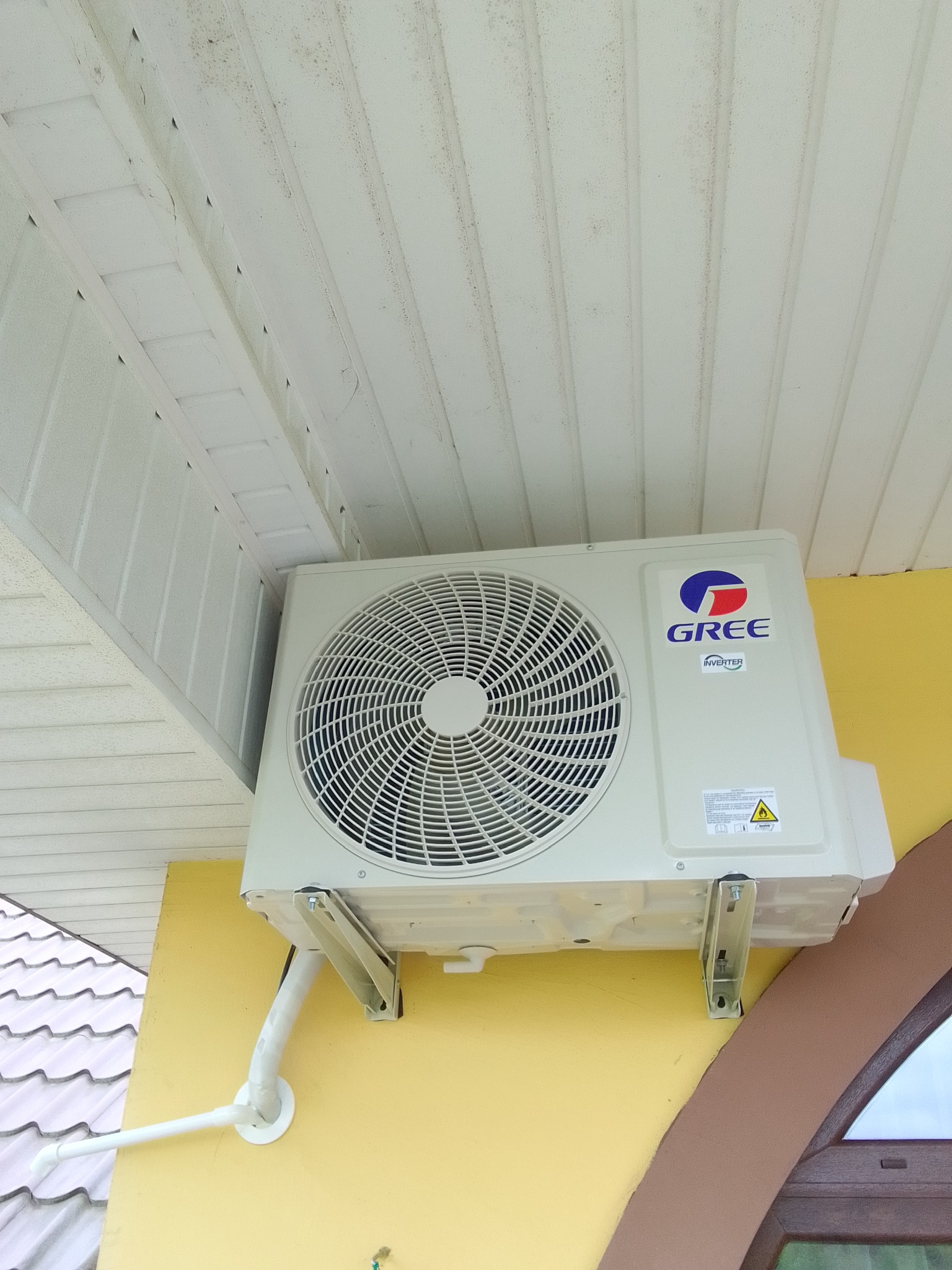 Klimatyzator Gree Inverter zamontowany na żółtej ścianie pod białym podbitkiem dachowym. Widoczne białe rury odprowadzające skropliny. Kąt ujęcia z dołu.