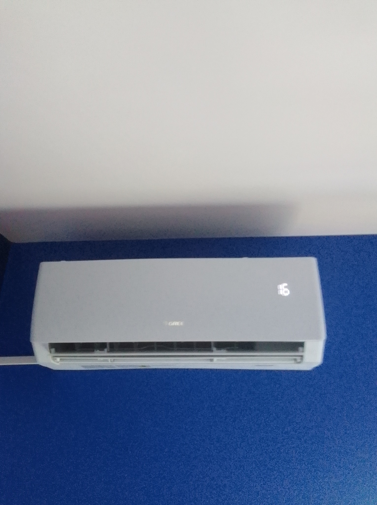Szary klimatyzator marki Gree z wyświetlaną temperaturą 16 stopni, zamontowany na niebieskiej ścianie pod białym sufitem.
