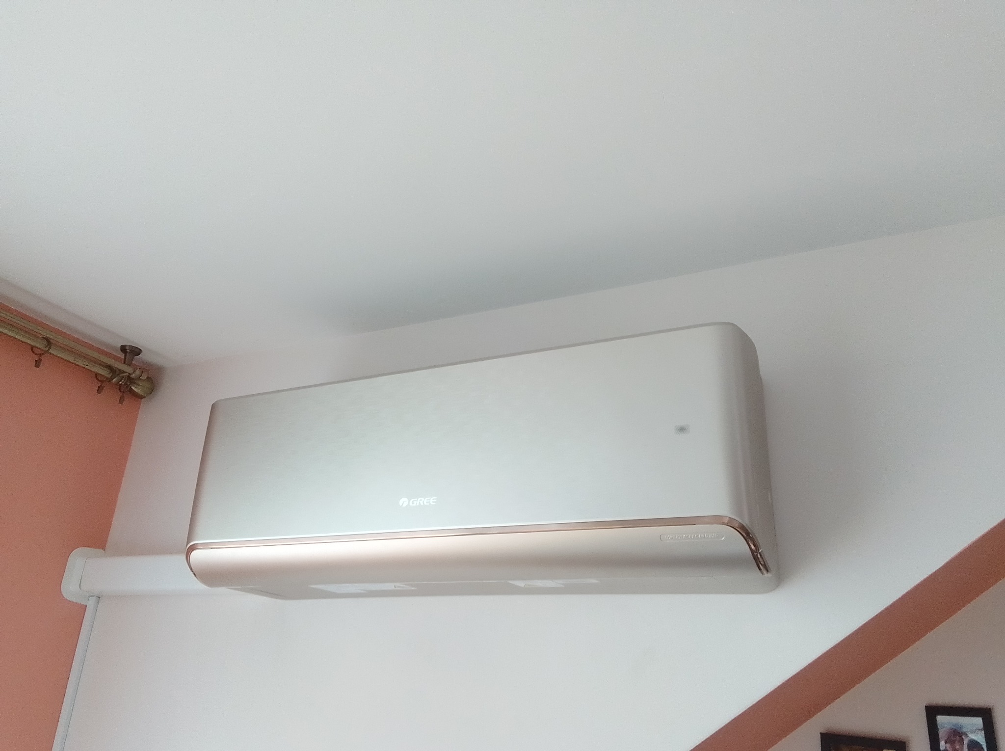 Klimatyzator Gree, model ścienny, zamontowany wewnątrz pomieszczenia. Widoczne detale montażu i wykończenia, biała obudowa z delikatnym, złotym akcentem.