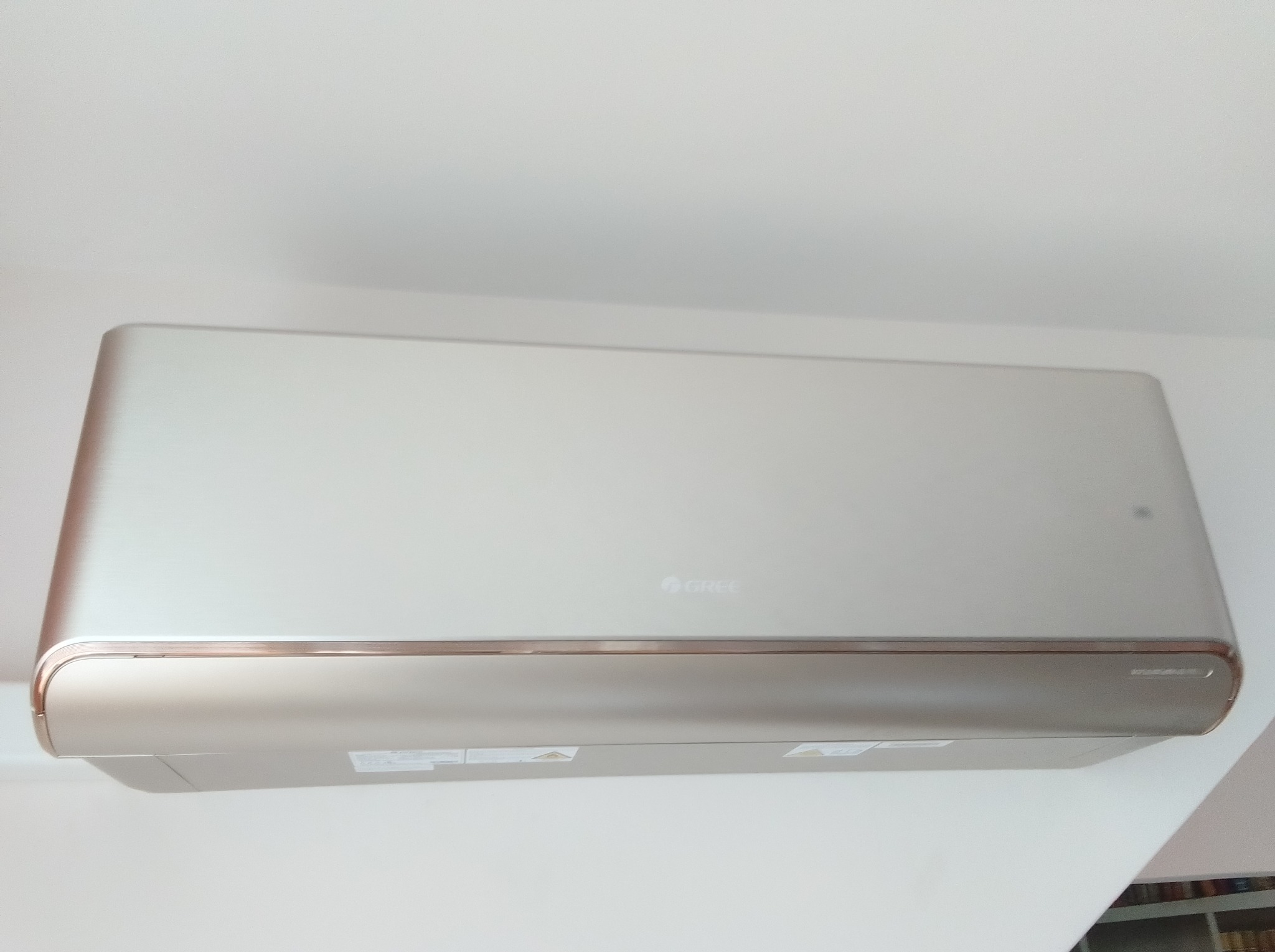 Elegancki klimatyzator marki Gree w odcieniach beżu i złota, zamontowany na białej ścianie. Widoczny fragment półki z książkami w tle.