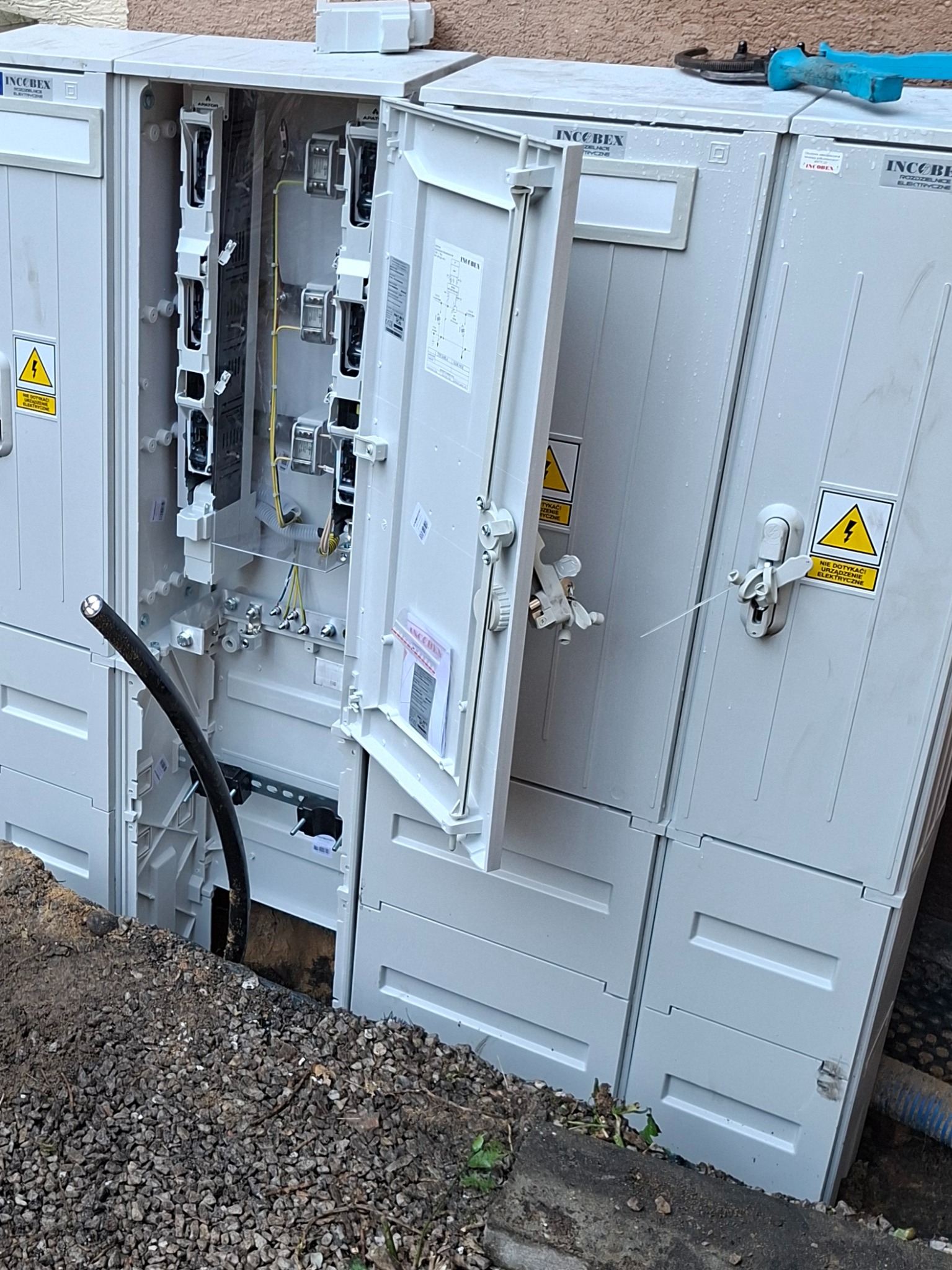 Otwarta skrzynka elektryczna IncoBex z kablem w ziemi, ostrzeżenie o napięciu. Widoczna instalacja wewnątrz. Na górze leży klucz do rur.