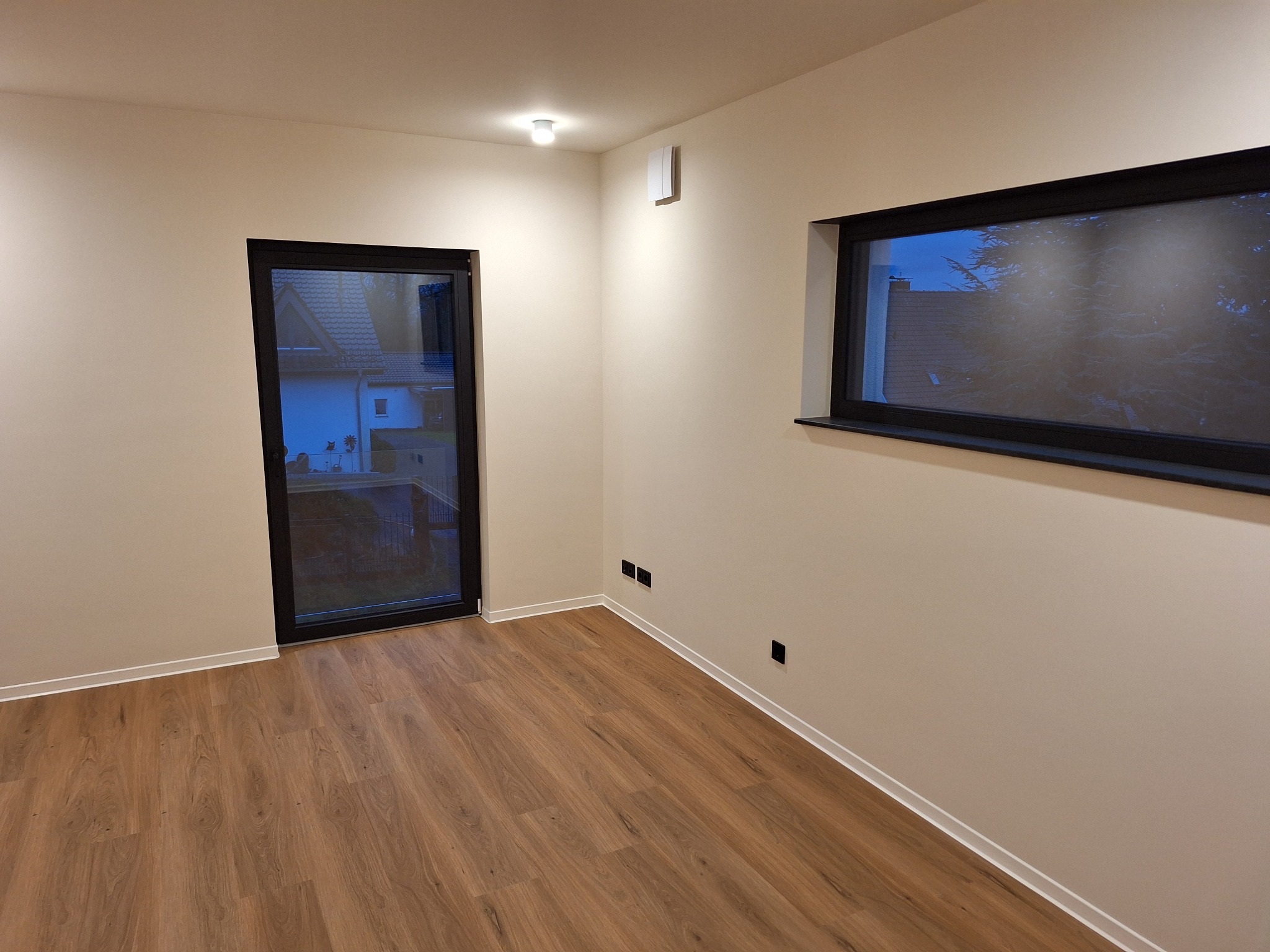 Pusty pokój po remoncie z panelami podłogowymi, świeżo pomalowanymi ścianami w odcieniach beżu, czarną stolarką okienną i drzwiową oraz widocznymi gniazdkami elektrycznymi.