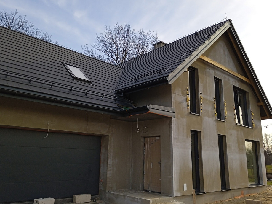 Surowy dom jednorodzinny z ciemną dachówką i grafitową bramą garażową. Widoczne ocieplenie wokół okien. Stan surowy zamknięty, bez tynków elewacyjnych.