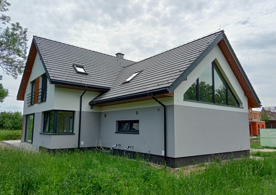 Nowoczesny dom z dwuspadowym dachem krytym szarą dachówką, z dużym trójkątnym oknem szczytowym i dwukolorową elewacją w odcieniach szarości. Widok z zewnątrz, trawa na pierwszym planie.