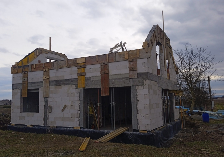 Surowy stan domu z białych bloczków, z widocznym szalunkiem na dachu i nadprożach. W tle pochmurne niebo i drzewo bez liści, ukazujące etap budowy domu murowanego.