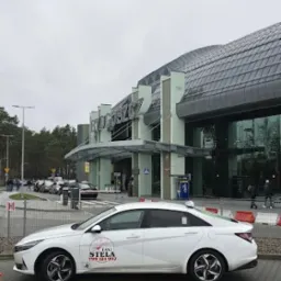 Biały samochód taxi z logo 'Taxi Stela' na tle budynku lotniska w Bydgoszczy, widok z boku.