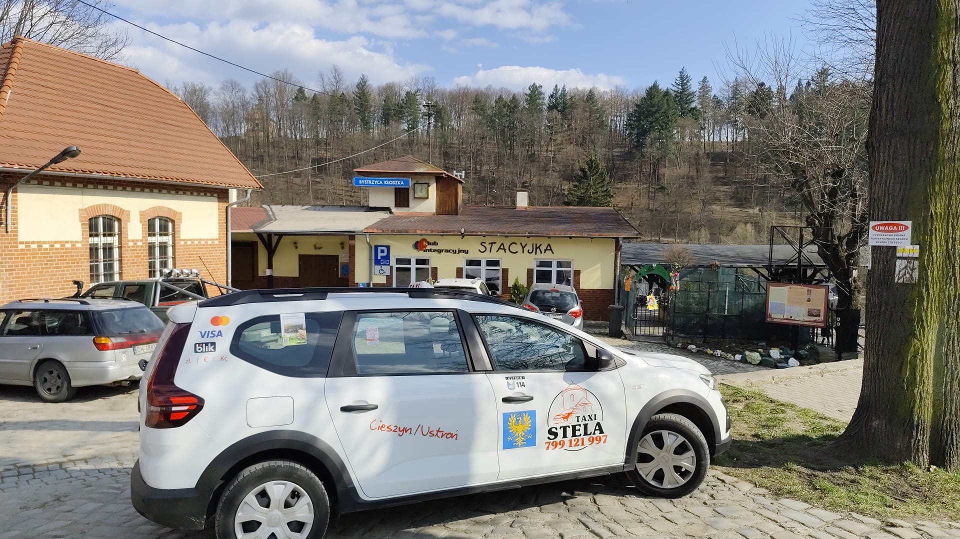 Biały samochód taxi z logo 'Taxi Stela' i oznaczeniami płatności VISA i BLIK zaparkowany przed budynkiem stacji kolejowej 'Bystrzyca Kłodzka'.