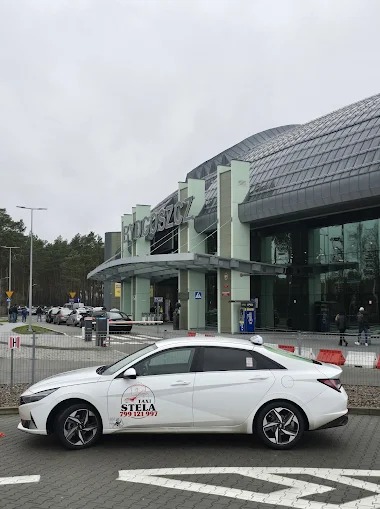 Biały samochód taxi z logo 'Taxi Stela' na tle budynku lotniska w Bydgoszczy, widok z boku.