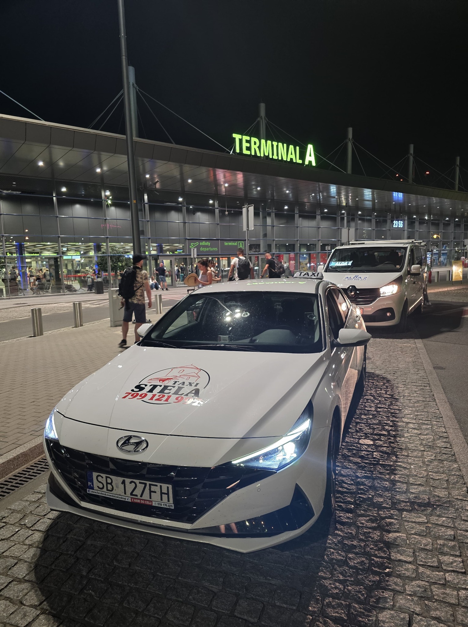 Biała taksówka z logo firmy 'STELA TAXI' na masce, zaparkowana przed terminalem A lotniska w nocy, widoczny drugi bus taksówkowy w tle i pasażerowie wchodzący do terminalu.