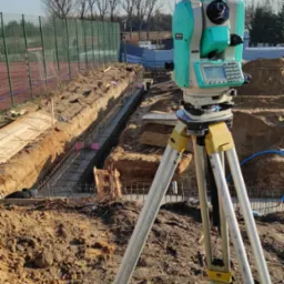 Zielony niwelator na statywie, ustawiony na tle wykopu zbrojonego pod fundamenty. Widoczne drewniane szalunki i stalowe pręty zbrojeniowe.