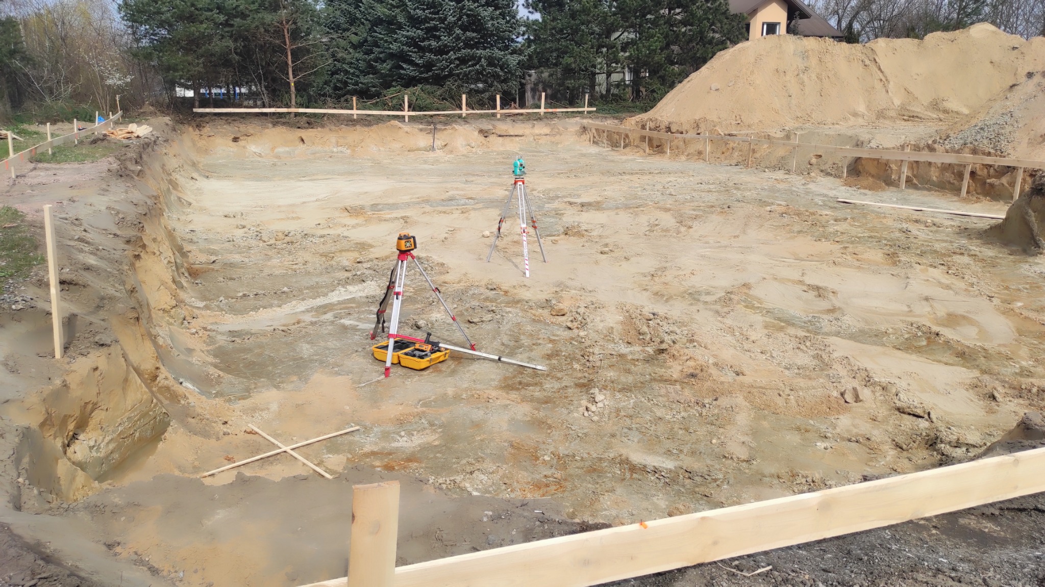Wykonany wykop pod fundamenty budynku, widoczne drewniane szalunki, niwelator laserowy i teodolit na statywach, oraz żółta skrzynka z narzędziami.