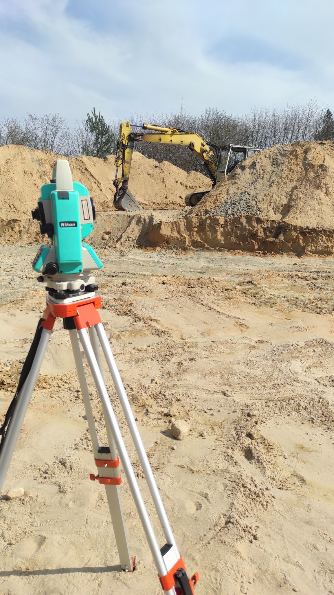Nikon Total Station na trójnogu, w tle wykop z żółtą koparką, teren budowy w piasku.