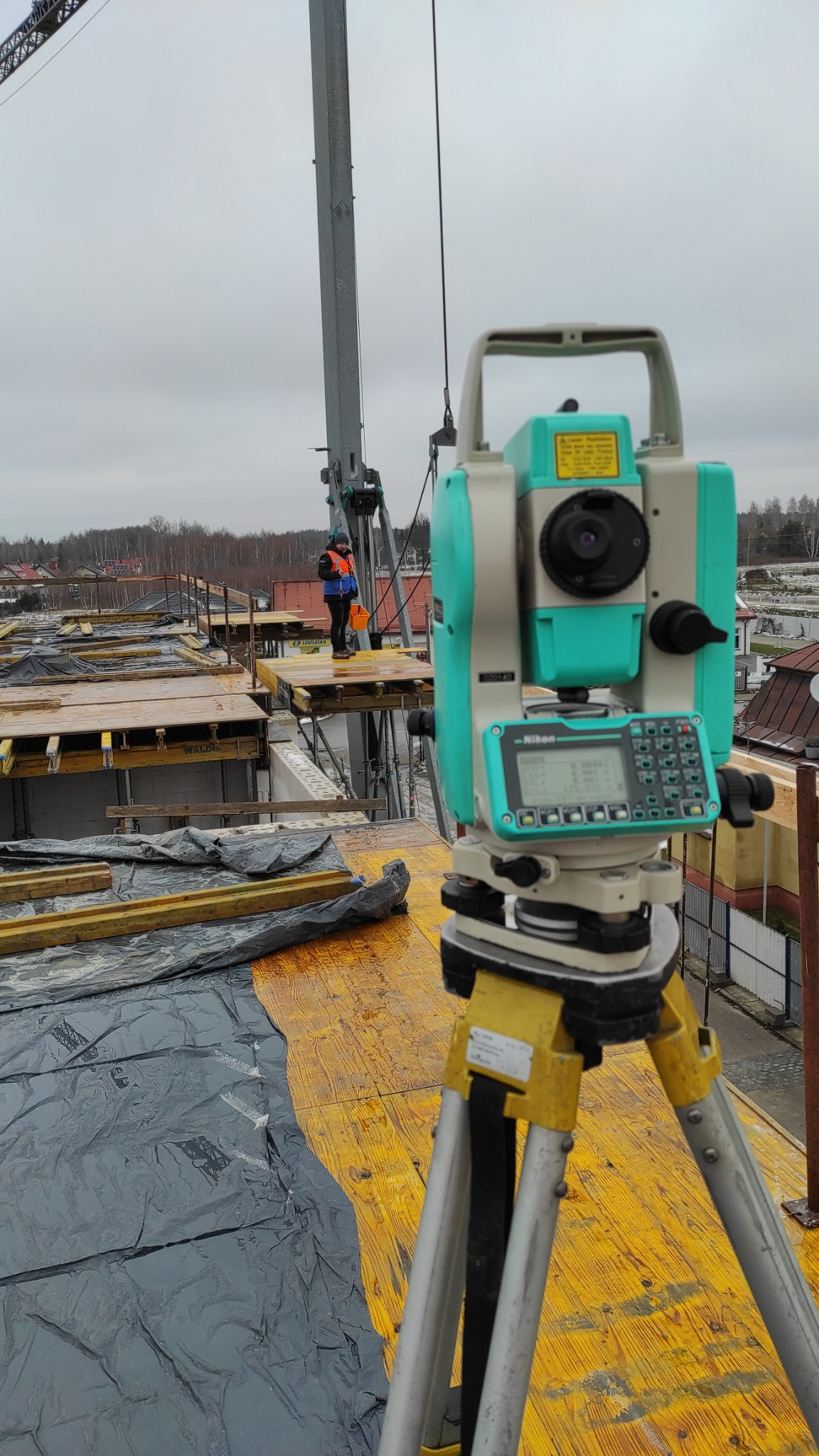 Nikon Total Station na statywie, ustawiony na budowie na tle pracującego operatora dźwigu i widocznych elementów szalunków stropowych.