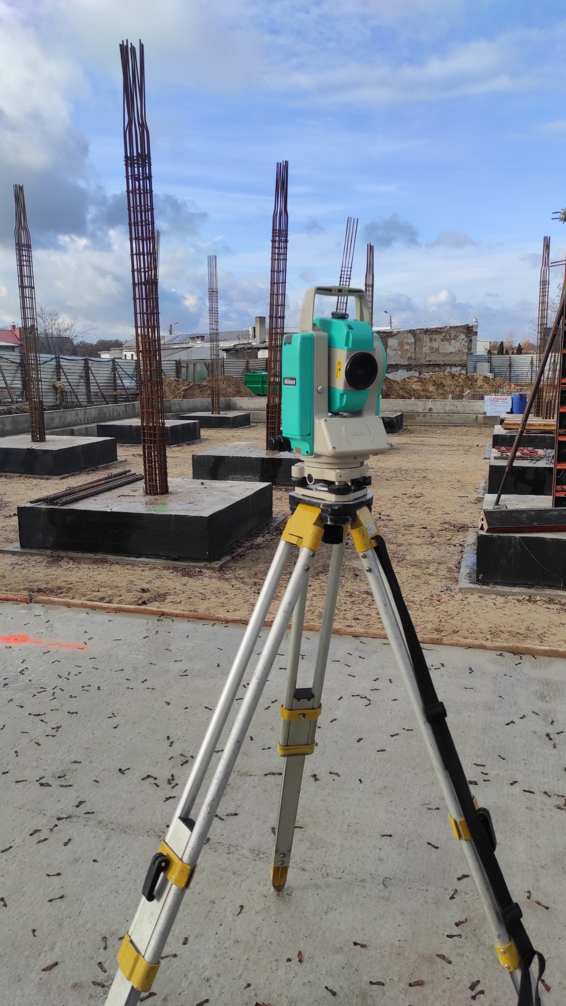 Nikon Total Station na trójnogu, zbrojenia fundamentów w tle. Widok na plac budowy z fundamentami i wystającymi prętami zbrojeniowymi.
