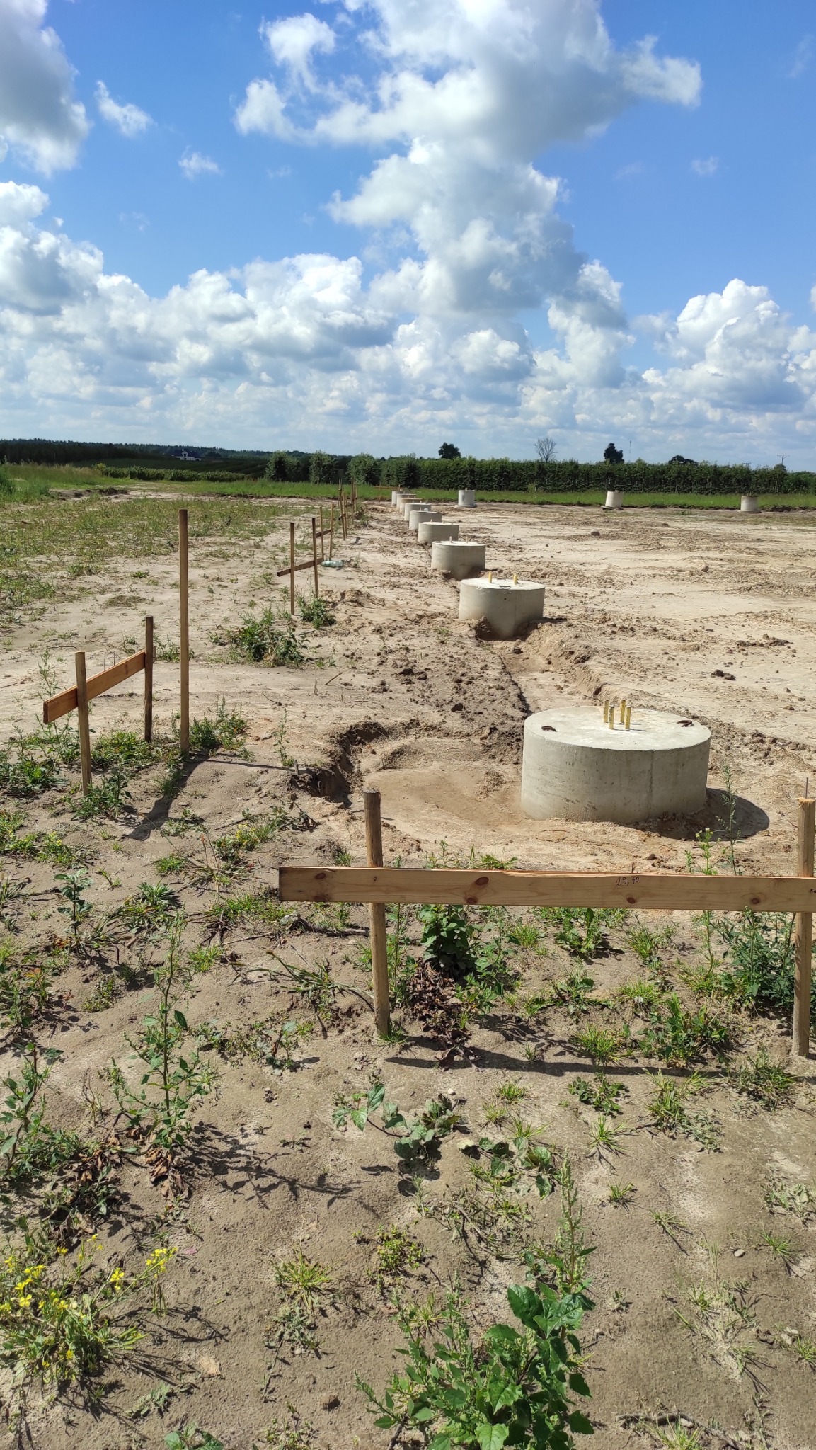 Wykonane fundamenty pod budowę, widoczne wystające pręty zbrojeniowe. Ustawione drewniane paliki geodezyjne wyznaczające granice działki. Błękitne niebo z chmurami w tle.