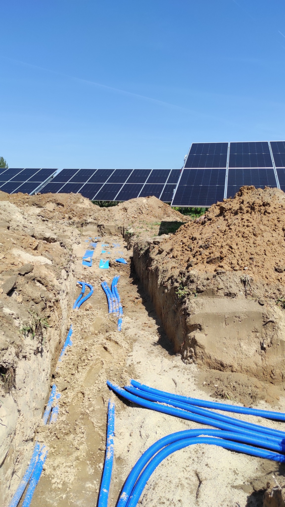 Instalacja rur w wykopie pod panele słoneczne, widok z góry, błękitne rury korugowane na tle piasku i ziemi, panele słoneczne w tle, błękitne niebo.