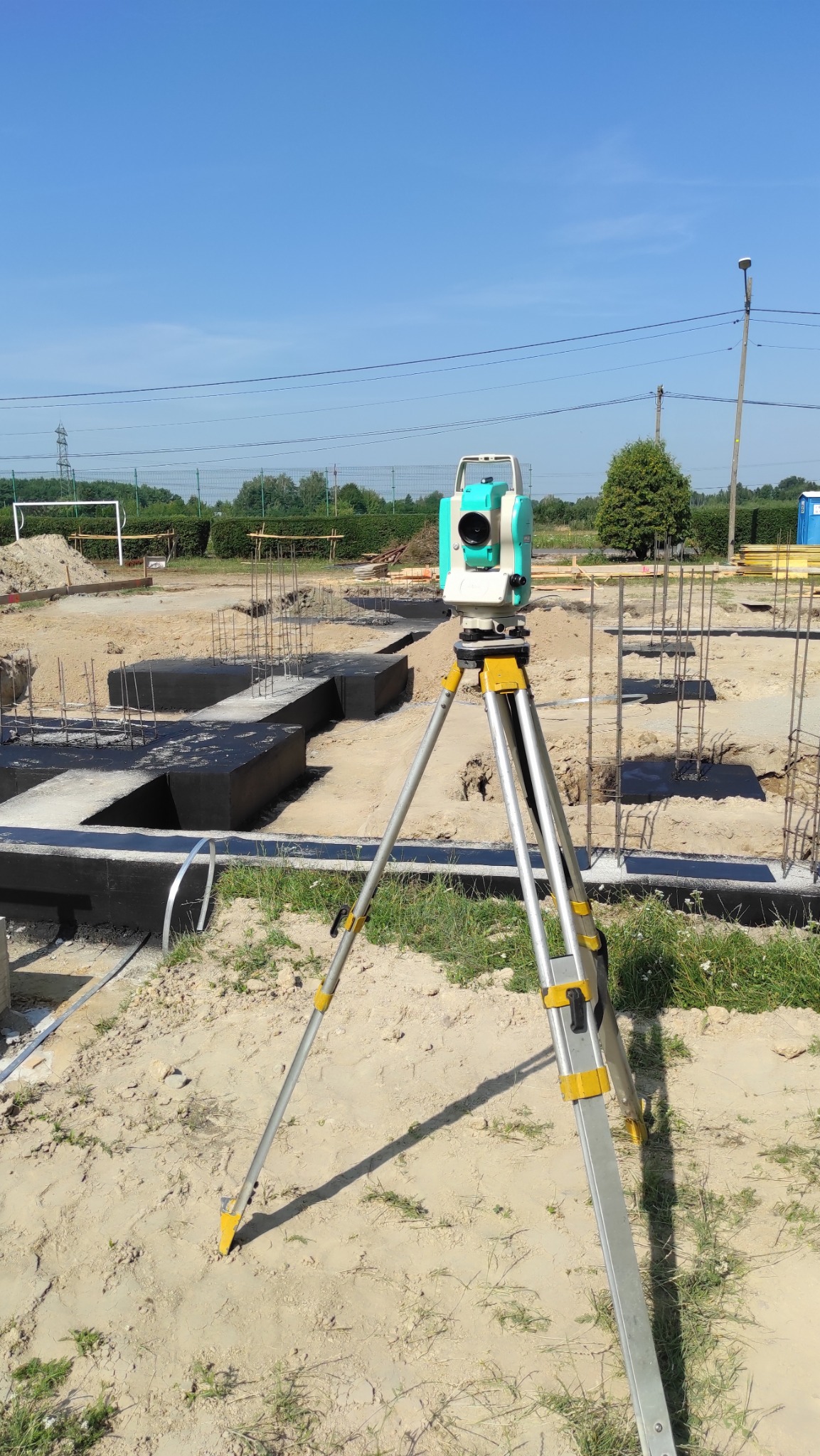Teodolit na statywie na placu budowy z widocznymi fundamentami i zbrojeniami. Sprzęt pomiarowy w trakcie pracy na tle błękitnego nieba i zieleni.