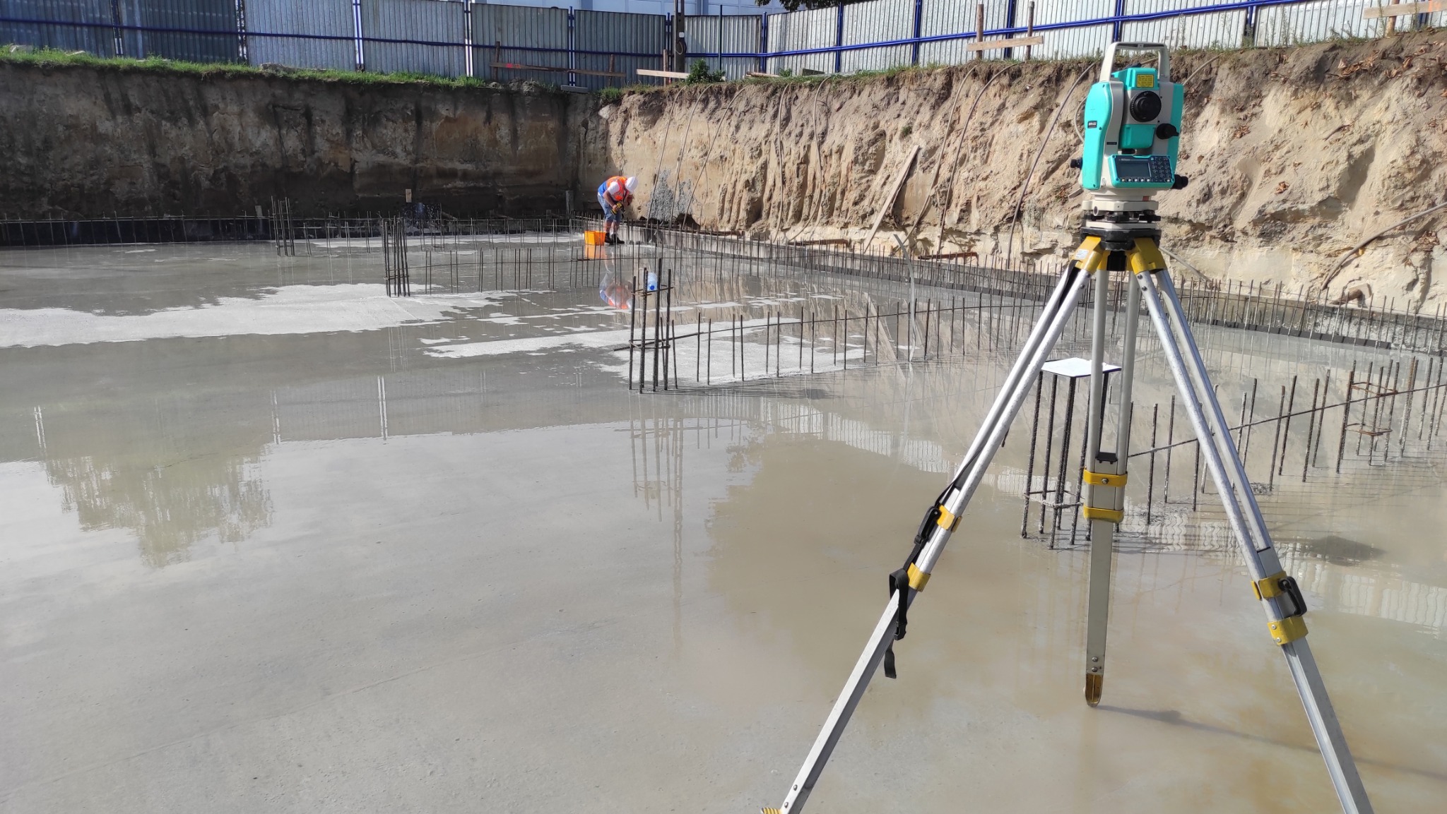 Niwelator na trójnogu na tle zalanego fundamentu zbrojonego; w tle pracownik budowlany. Widok na plac budowy z bliska, z naciskiem na pomiar geodezyjny.