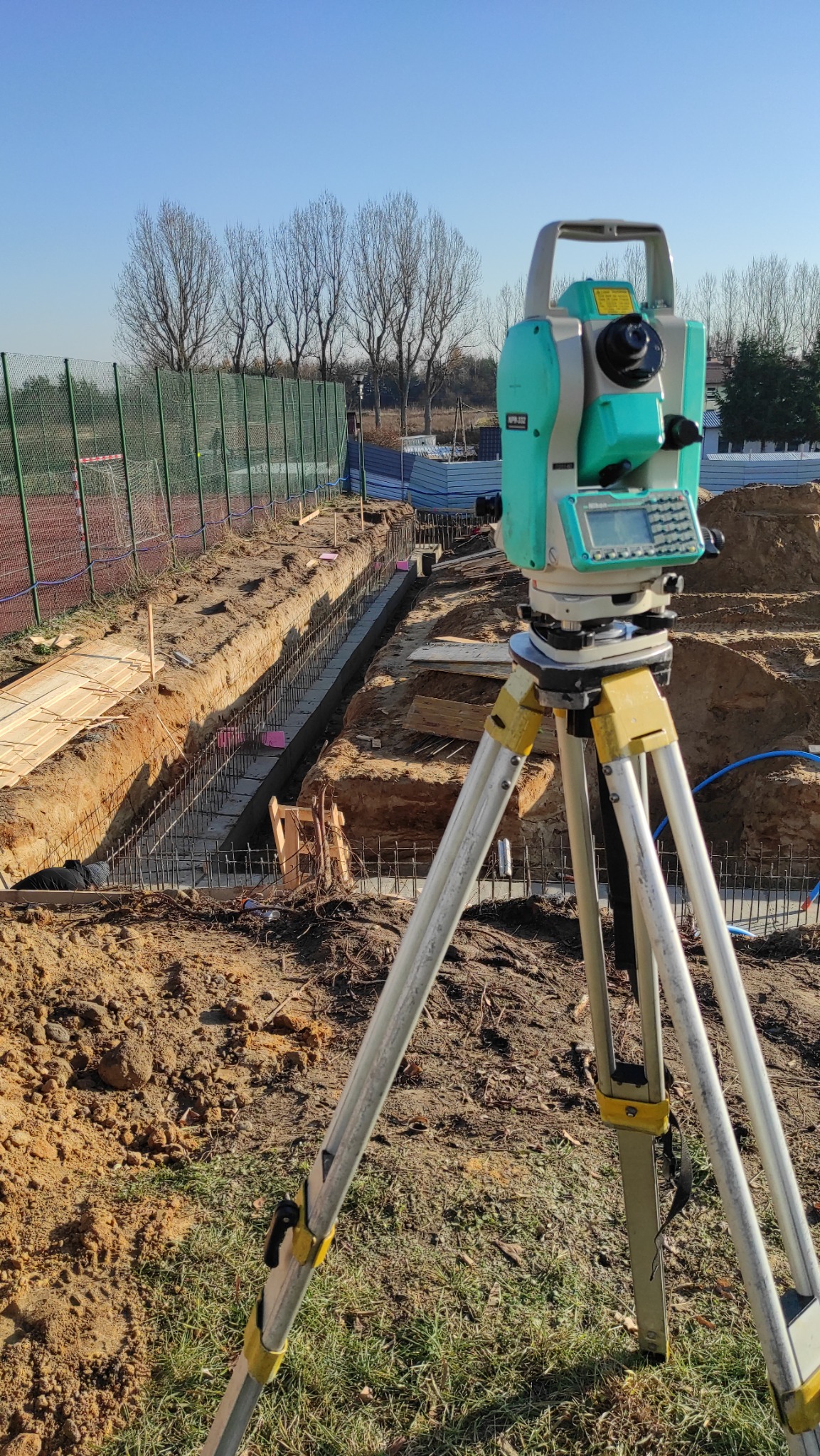 Zielony niwelator na statywie, ustawiony na tle wykopu zbrojonego pod fundamenty. Widoczne drewniane szalunki i stalowe pręty zbrojeniowe.