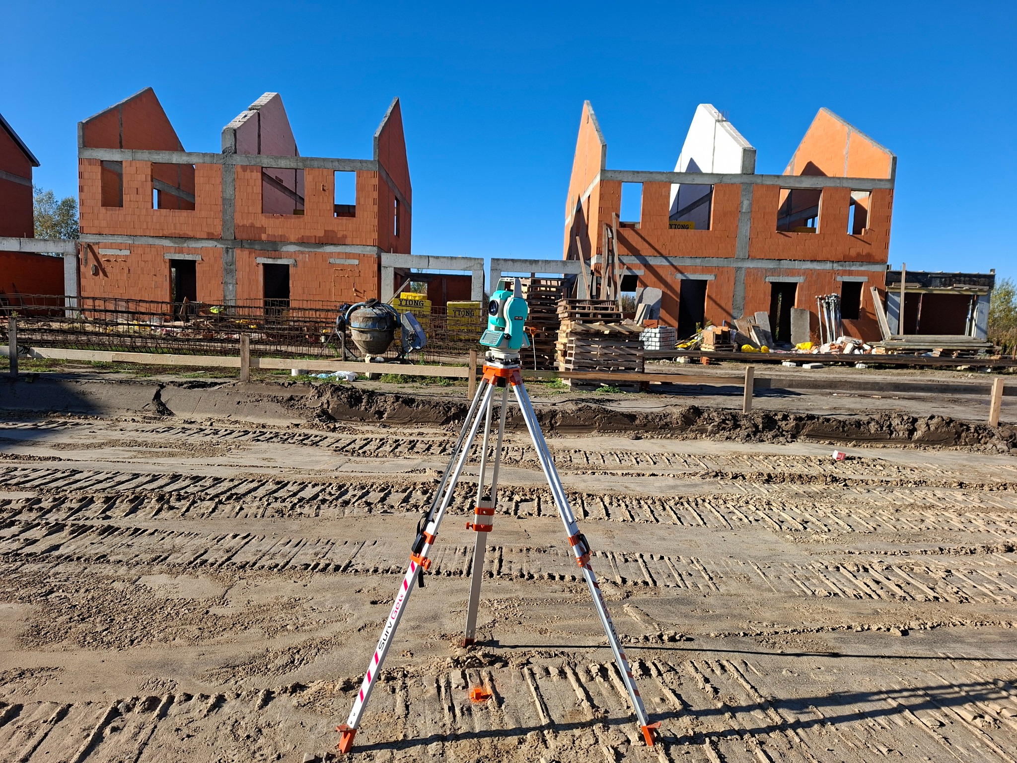 Niwelator na statywie na placu budowy z dwoma budynkami w stanie surowym otwartym w tle, w słoneczny dzień. Widoczne ślady opon na piasku.