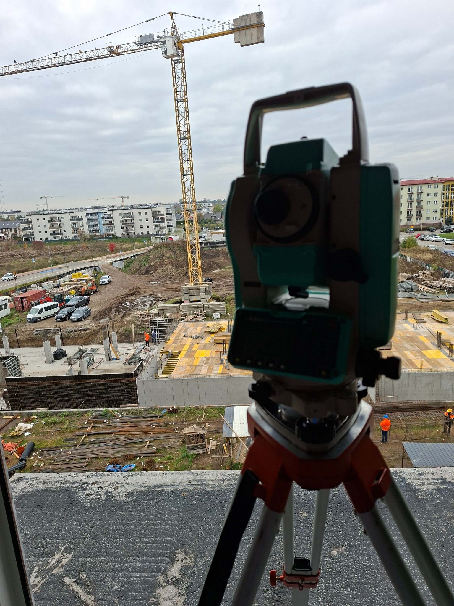 Teodolit na statywie na tle budowy z dźwigiem. Widok z góry na teren budowy, gdzie widać fundamenty i pracowników w pomarańczowych kamizelkach.