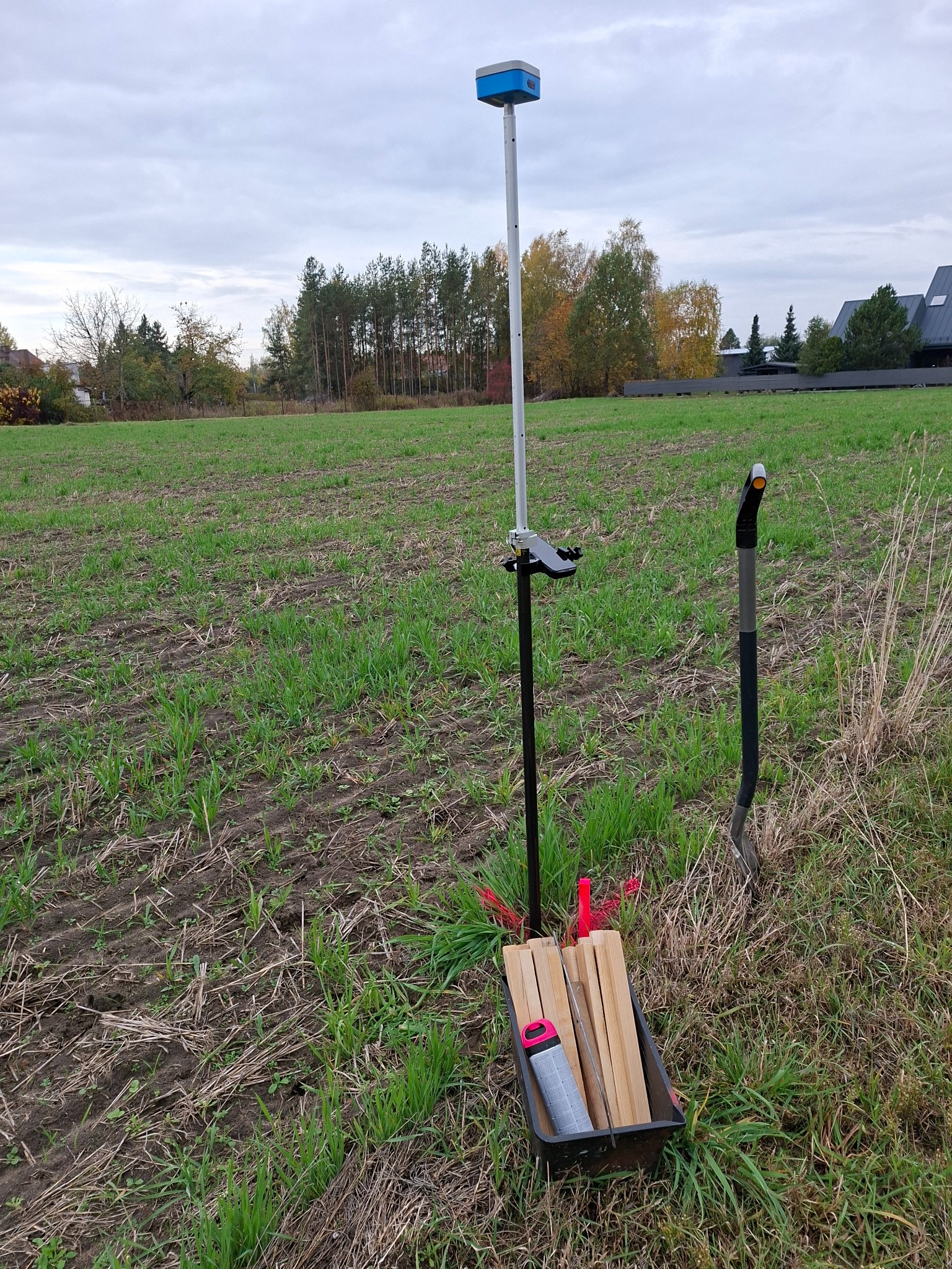 Pomiar geodezyjny w terenie: tyczka z odbiornikiem GPS, paliki, szpadel. Pole z młodą trawą, w tle drzewa i zabudowania. Pochmurne niebo.