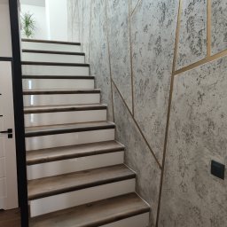 Witold Wdowiak ART.HOME Usługi Remontowe Budowlane - Schody z drewnianymi stopniami i białymi podstopnicami, ściana pokryta dekoracyjnym tynkiem w odcieniach szarości z geometrycznymi, złotymi wstawkami. Widoczny fragment białych drzwi z czarną klamką.