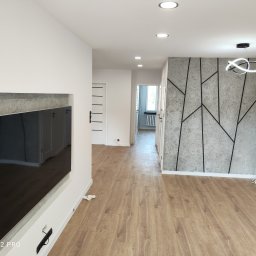 Witold Wdowiak ART.HOME Usługi Remontowe Budowlane - Nowoczesny salon z drewnianą podłogą, ścianą akcentową z betonowym wzorem geometrycznym, wnęką na telewizor i designerską lampą sufitową.