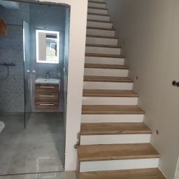 Witold Wdowiak ART.HOME Usługi Remontowe Budowlane - Wnętrze w trakcie prac: Schody z drewnianymi stopniami i białymi podstopnicami, obok wejście do łazienki z szarą glazurą, umywalką z szafką z drewnopodobnym fornirem i lustrem z podświetleniem LED.