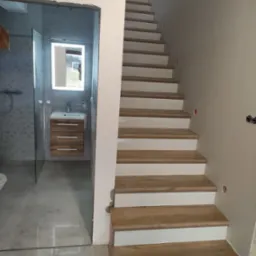 Wnętrze w trakcie prac: Schody z drewnianymi stopniami i białymi podstopnicami, obok wejście do łazienki z szarą glazurą, umywalką z szafką z drewnopodobnym fornirem i lustrem z podświetleniem LED.