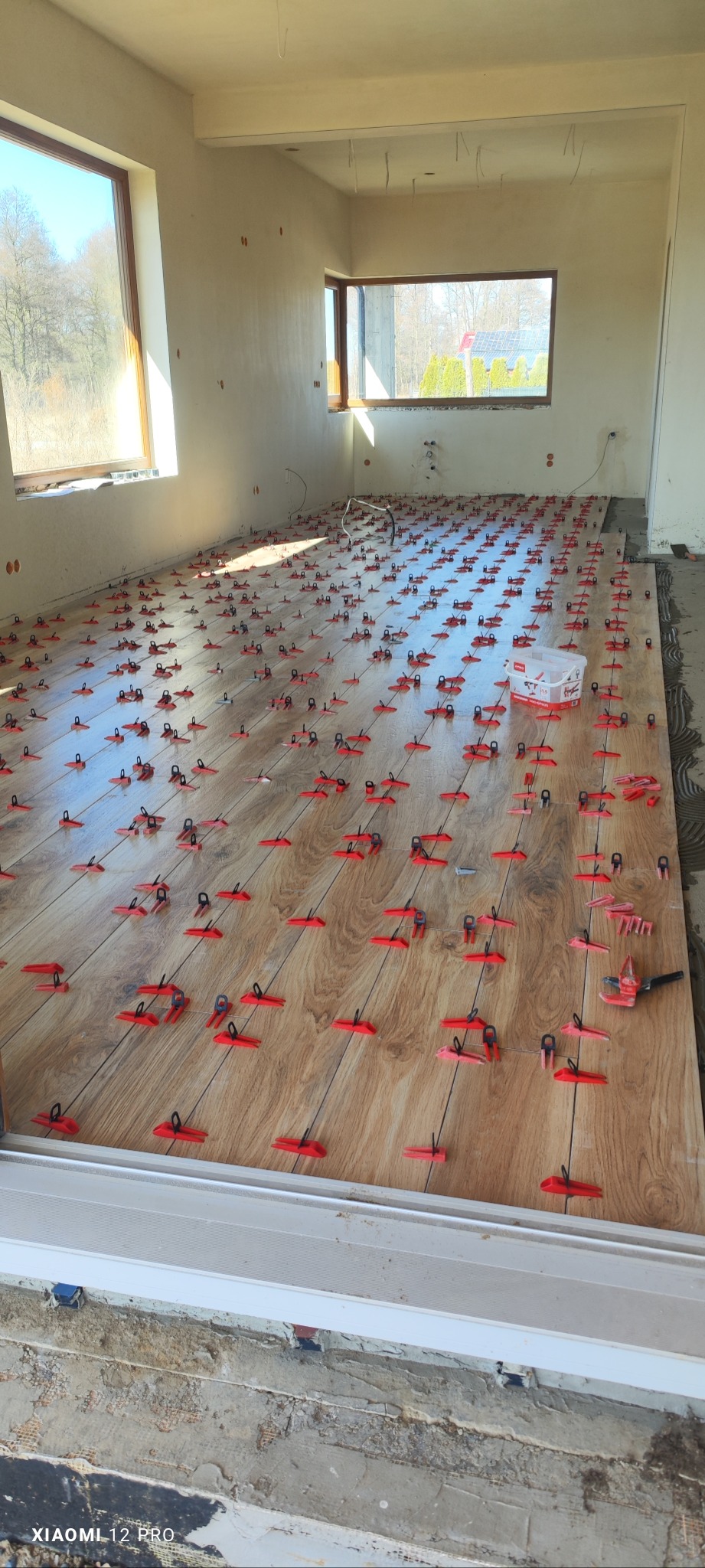 Układanie płytek podłogowych imitujących drewno z użyciem czerwonych klipsów poziomujących w jasnym, surowym pomieszczeniu z dużymi oknami.