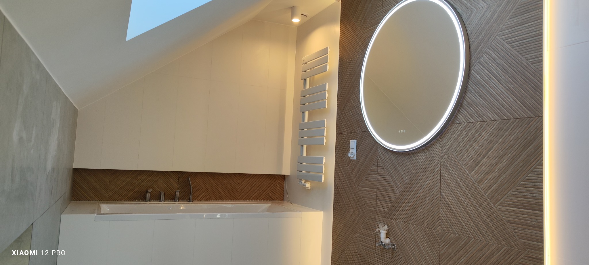 Nowoczesna łazienka na poddaszu z wanną obudowaną białą zabudową, grzejnikiem drabinkowym i okrągłym lustrem z podświetleniem LED na ścianie wyłożonej płytkami w geometryczny wzór.