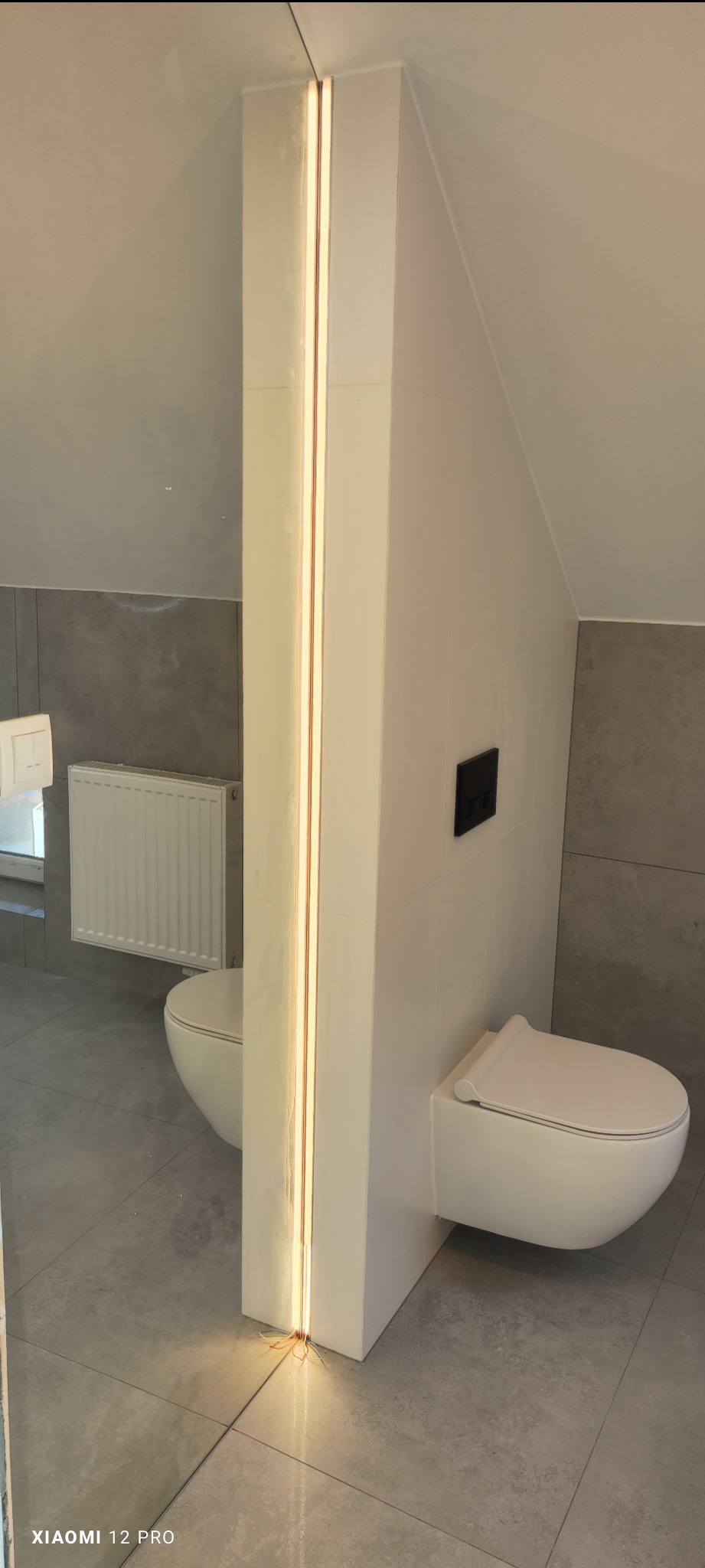 Nowoczesna łazienka z podwieszaną toaletą, szarą płytką i pionowym oświetleniem LED w szczelinie ściany działowej, odbijająca się w lustrze.