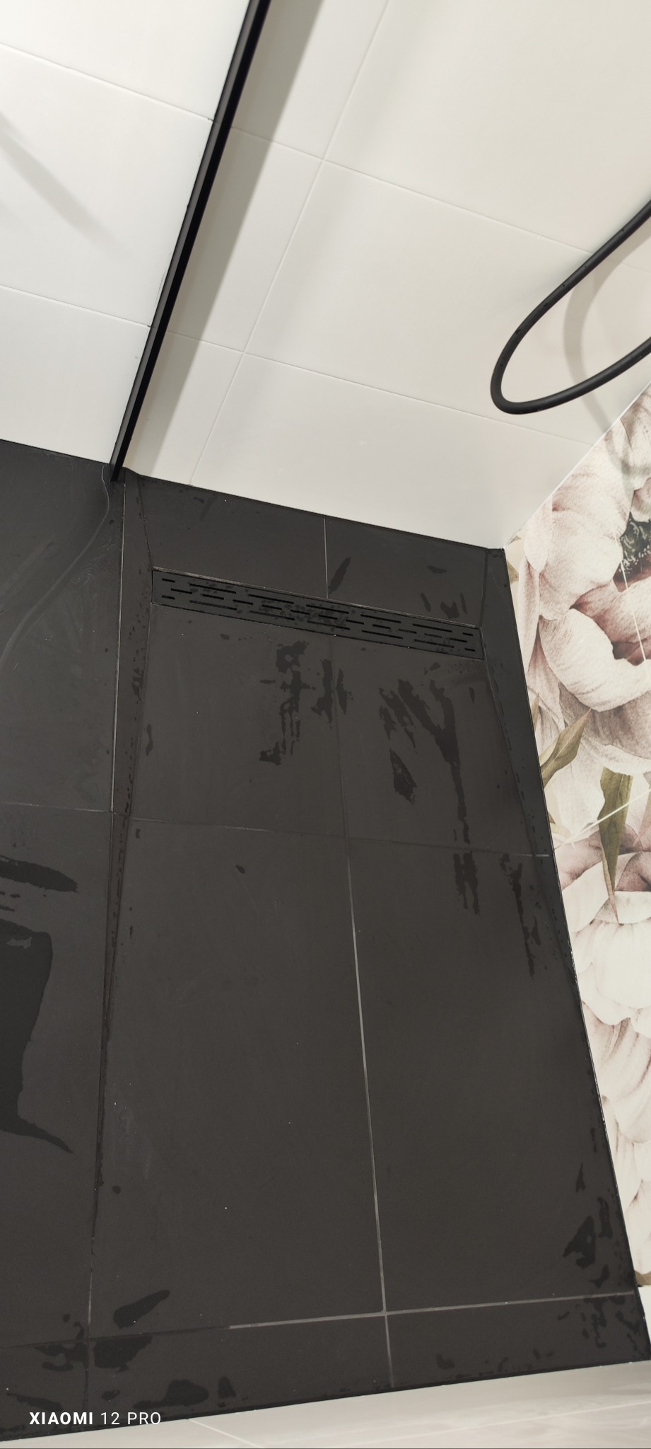 Ukończona instalacja liniowego odpływu prysznicowego w czarnych, błyszczących płytkach, z widocznymi śladami wody po testach szczelności, białe płytki na ścianie i dekoracyjna tapeta z motywem...