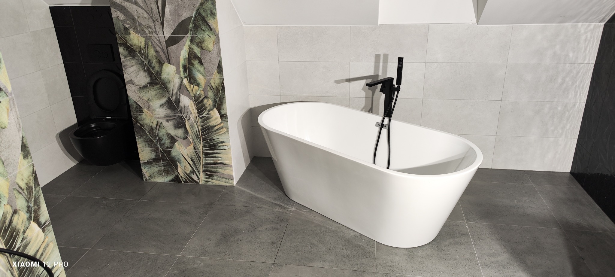 Nowoczesna łazienka z wolnostojącą białą wanną i czarną armaturą, szare płytki podłogowe i ścienne, akcent tropikalny na płytkach przy czarnej toalecie podtynkowej.
