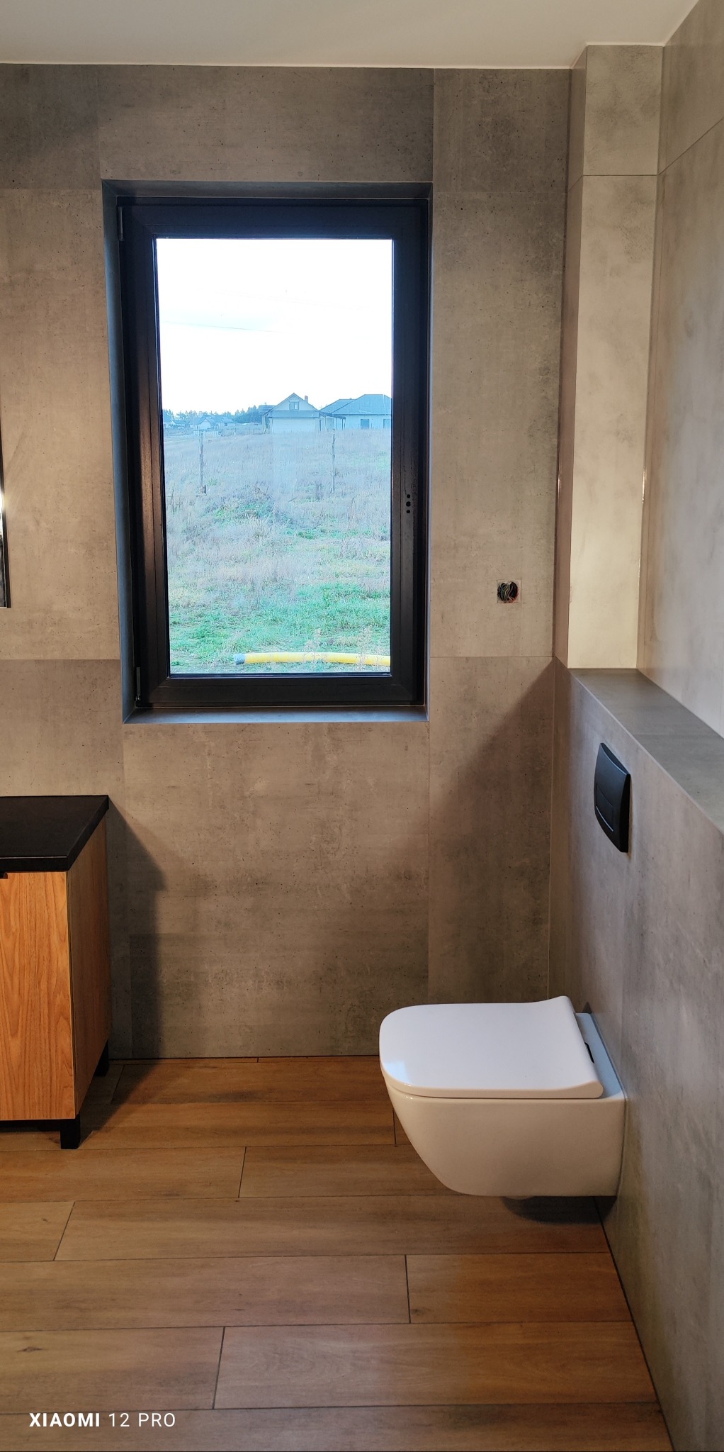 Nowoczesna łazienka z podwieszaną toaletą, drewnopodobną podłogą, szarą okładziną ścienną imitującą beton i oknem z widokiem na wiejski krajobraz.