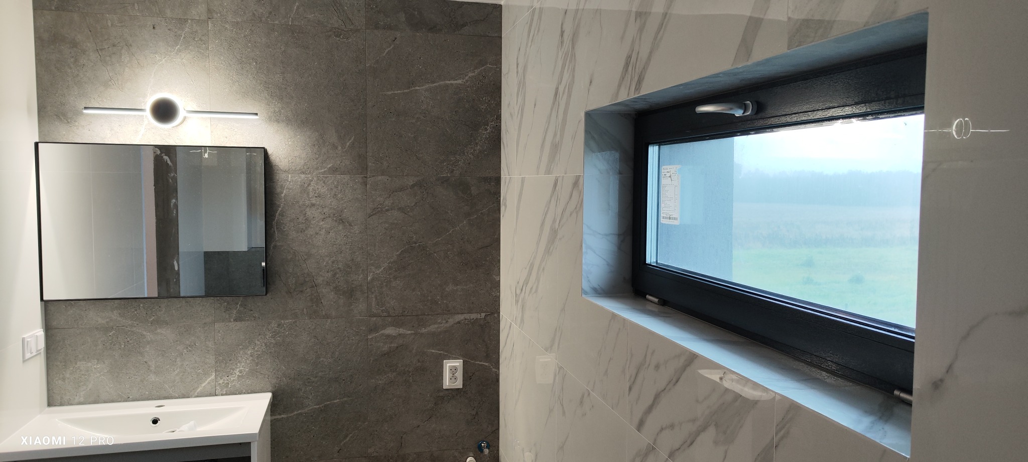 Nowoczesna łazienka z szarymi i białymi płytkami imitującymi marmur, czarnym oknem z widokiem na zewnątrz i podświetlanym lustrem nad umywalką.