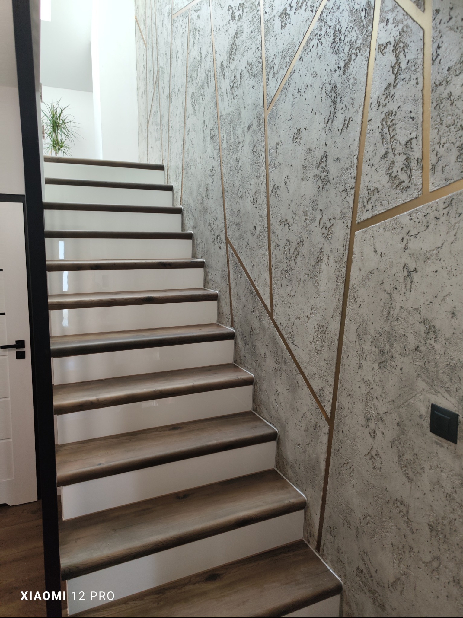 Schody z drewnianymi stopniami i białymi podstopnicami, ściana pokryta dekoracyjnym tynkiem w odcieniach szarości z geometrycznymi, złotymi wstawkami. Widoczny fragment białych drzwi z czarną klamką.