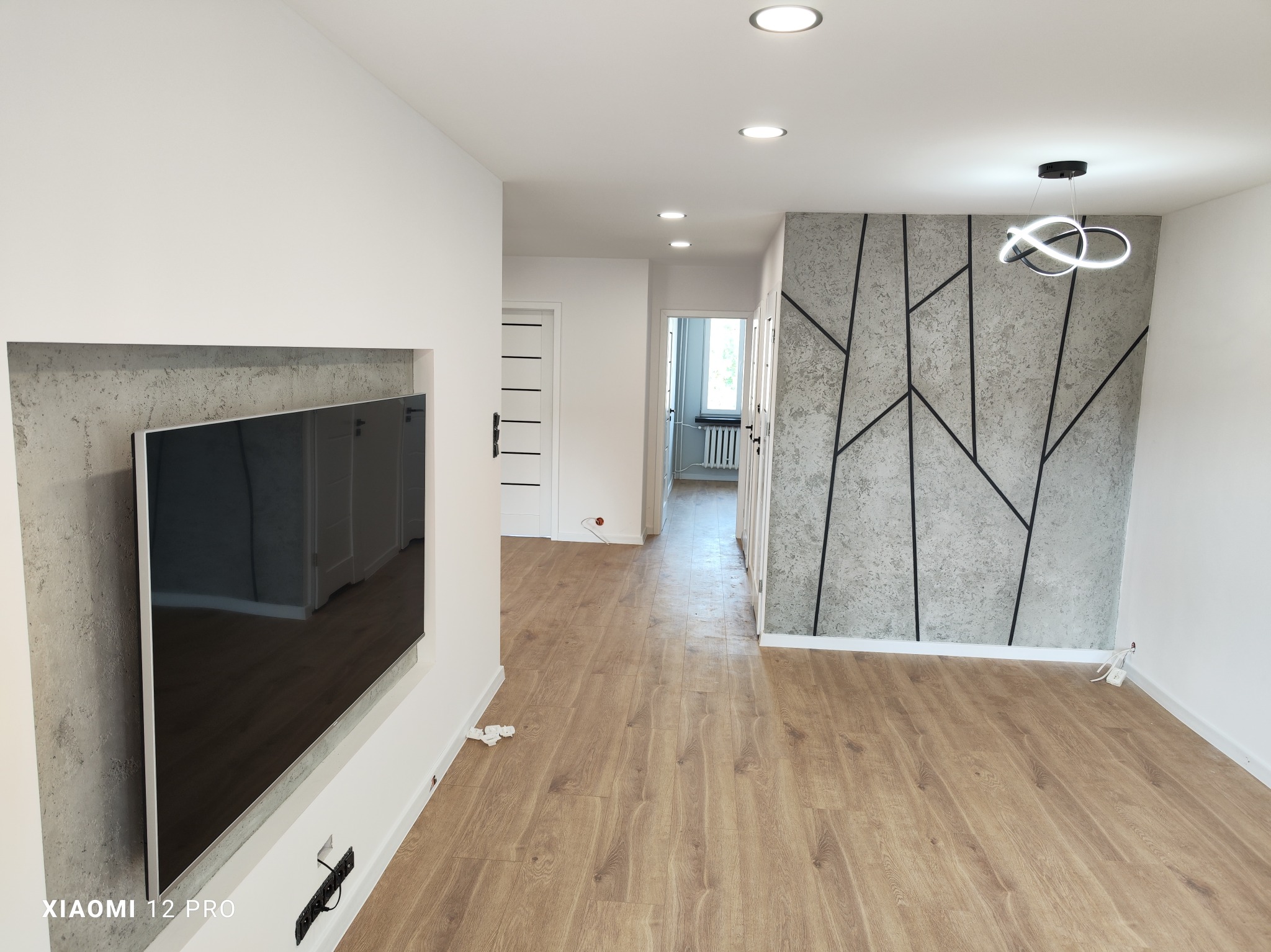 Nowoczesny salon z drewnianą podłogą, ścianą akcentową z betonowym wzorem geometrycznym, wnęką na telewizor i designerską lampą sufitową.