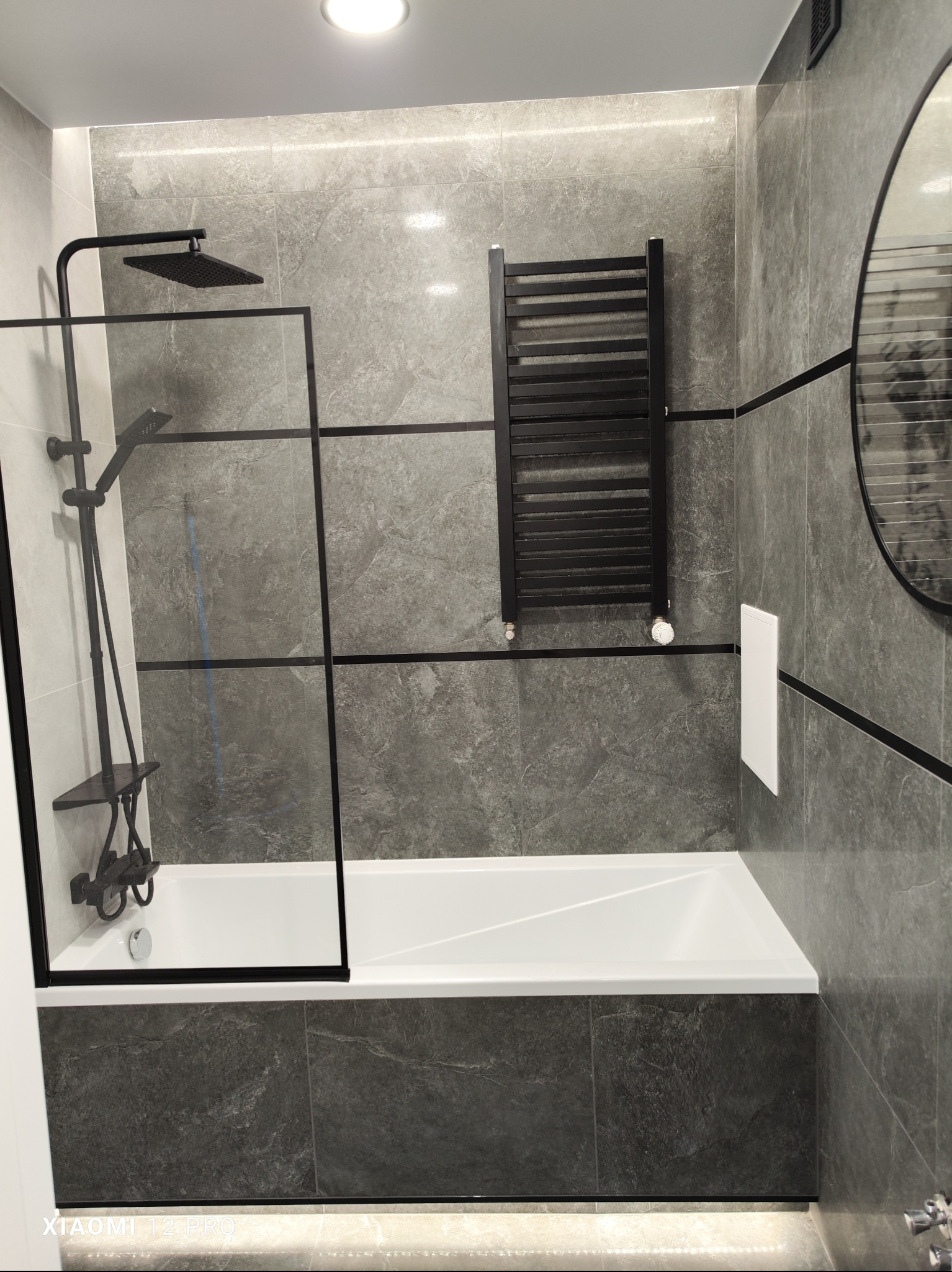Nowoczesna łazienka z szarymi płytkami imitującymi kamień, białą wanną z czarnym parawanem nawannowym, czarną armaturą prysznicową i grzejnikiem drabinkowym w tym samym kolorze. Okrągłe lustro...